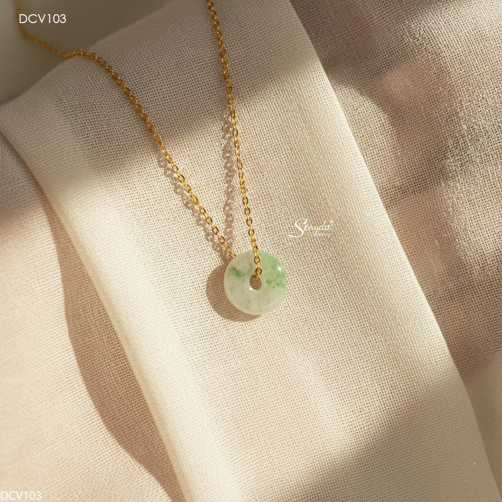 A delicate gold chain necklace with a round light green jade pendant, draped elegantly on a textured beige fabric background with soft, warm sunlight and shadows.