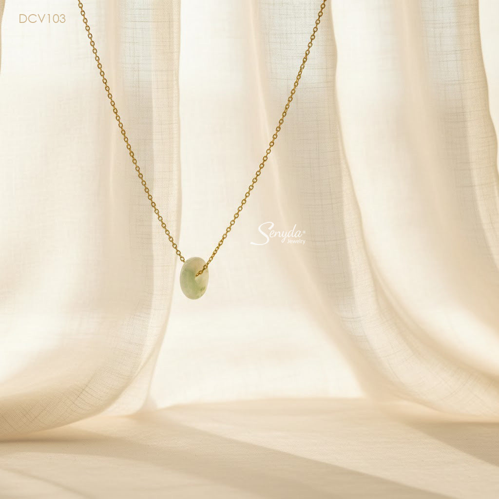 A delicate gold chain necklace with a small oval light green jade pendant, draped against a soft, cream-colored fabric backdrop with warm sunlight filtering through.