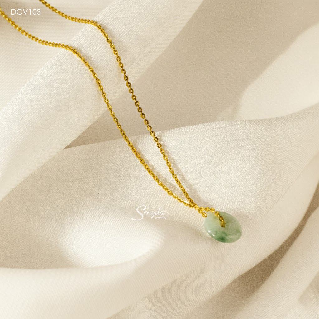 A gold chain necklace with a small, polished light green jadeite pendant, displayed on draped cream-colored fabric.