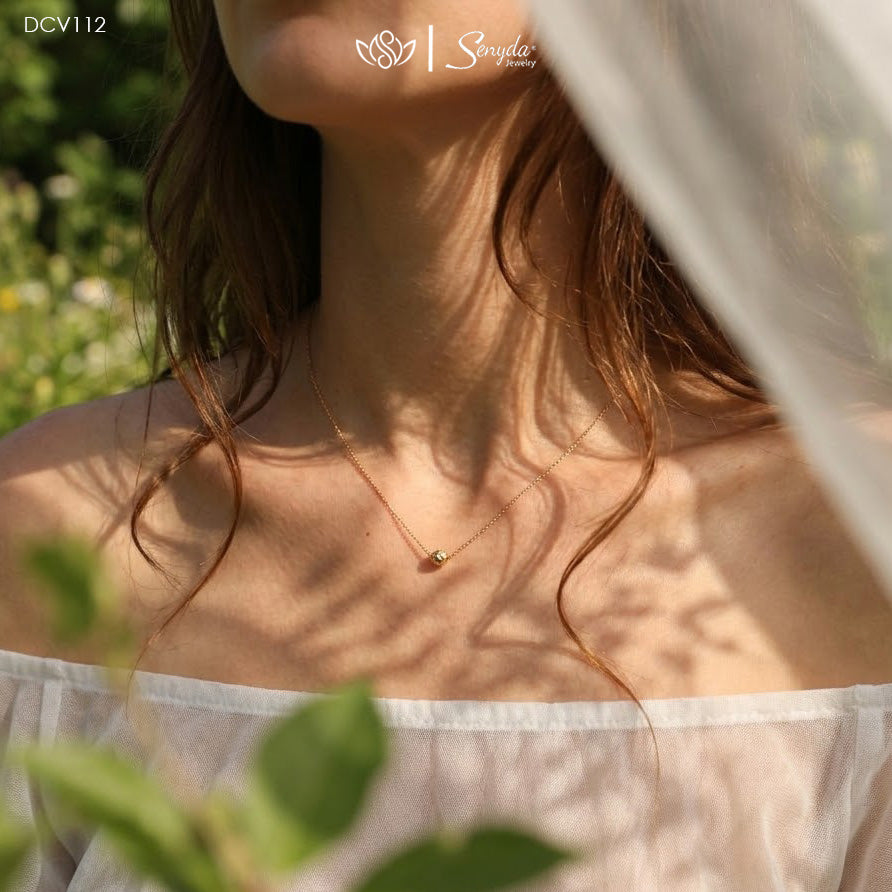 A delicate gold chain necklace with a small gold bead pendant worn by a woman outdoors. The image features dappled sunlight and shadows on her skin, with a white veil in the foreground and green foliage in the background.
