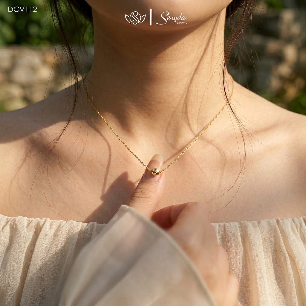A delicate gold chain necklace with a small gold bead pendant worn by a model. The shot is taken outdoors in warm sunlight, highlighting the fine jewelry on the model's neck, with a soft beige off-the-shoulder top visible.
