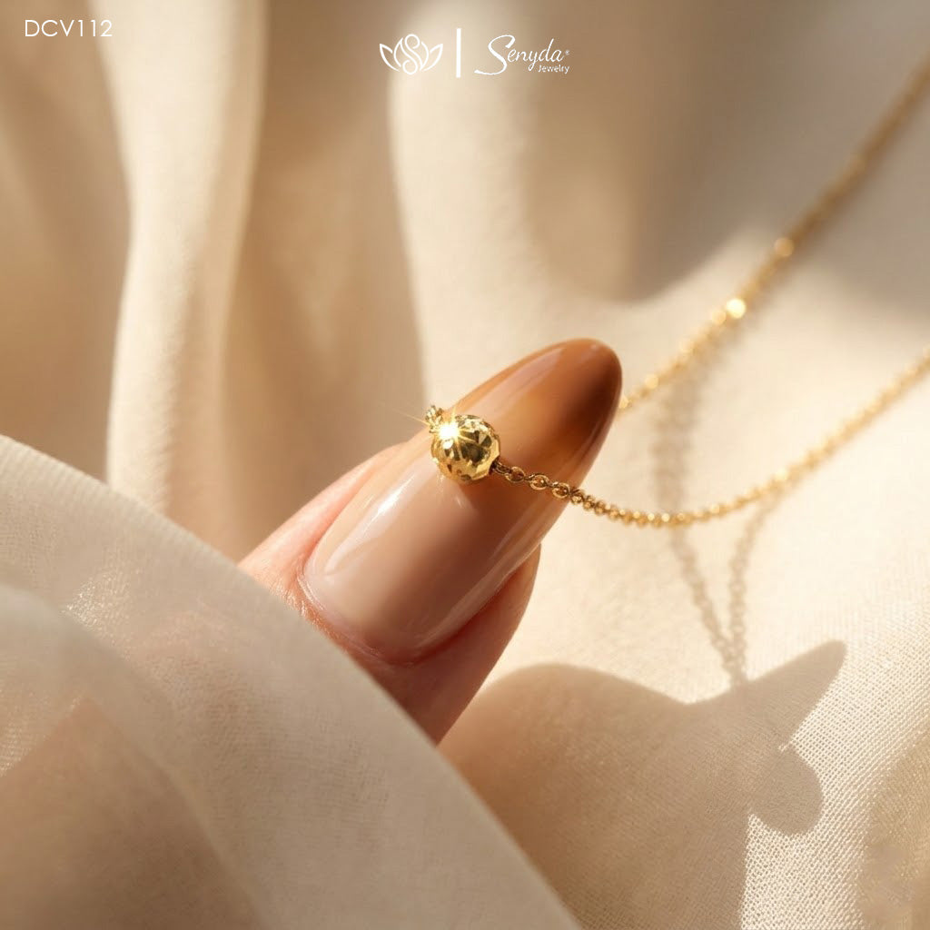 A close-up lifestyle shot of a delicate gold chain necklace with a small gold bead pendant, held by a manicured finger against a soft beige fabric backdrop. The brand logo 'Senya Gold' is visible at the top.