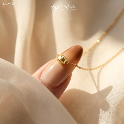 A close-up lifestyle shot of a delicate gold chain necklace with a small gold bead pendant, held by a manicured finger against a soft beige fabric backdrop. The brand logo 'Senya Gold' is visible at the top.