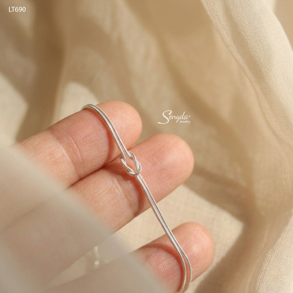 Sterling silver double snake chain bracelet tied in a love knot, shown flat on a cream background