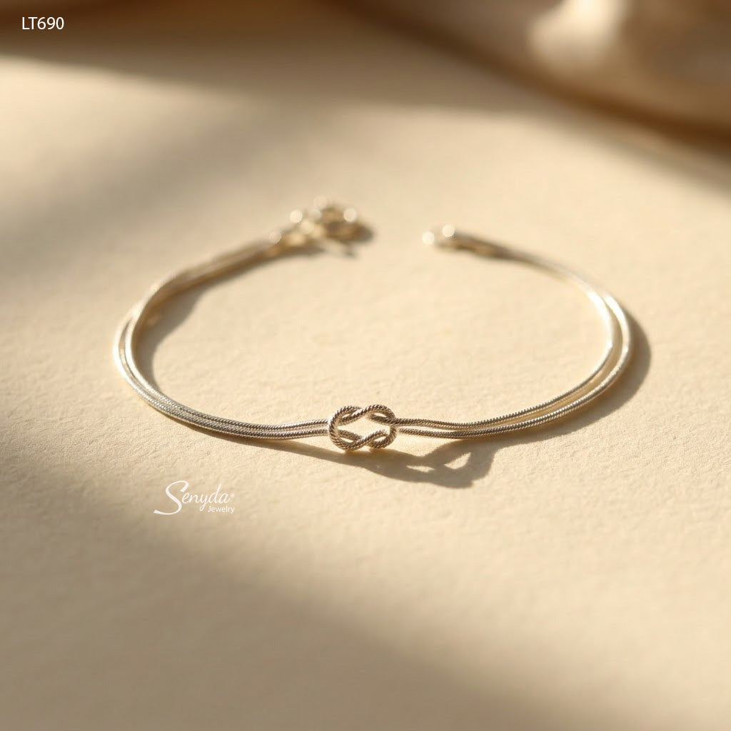 Sterling silver double snake chain bracelet tied in a love knot, shown flat on a cream background