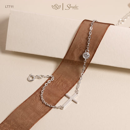 A silver chain bracelet with a small cross charm and a round clear stone, draped over a brown sheer ribbon on a cream-colored surface. The brand name 'Sonya' is visible in the top right corner.