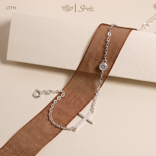 A silver chain bracelet with a small cross charm and a round clear stone, draped over a brown sheer ribbon on a cream-colored surface. The brand name 'Sonya' is visible in the top right corner.