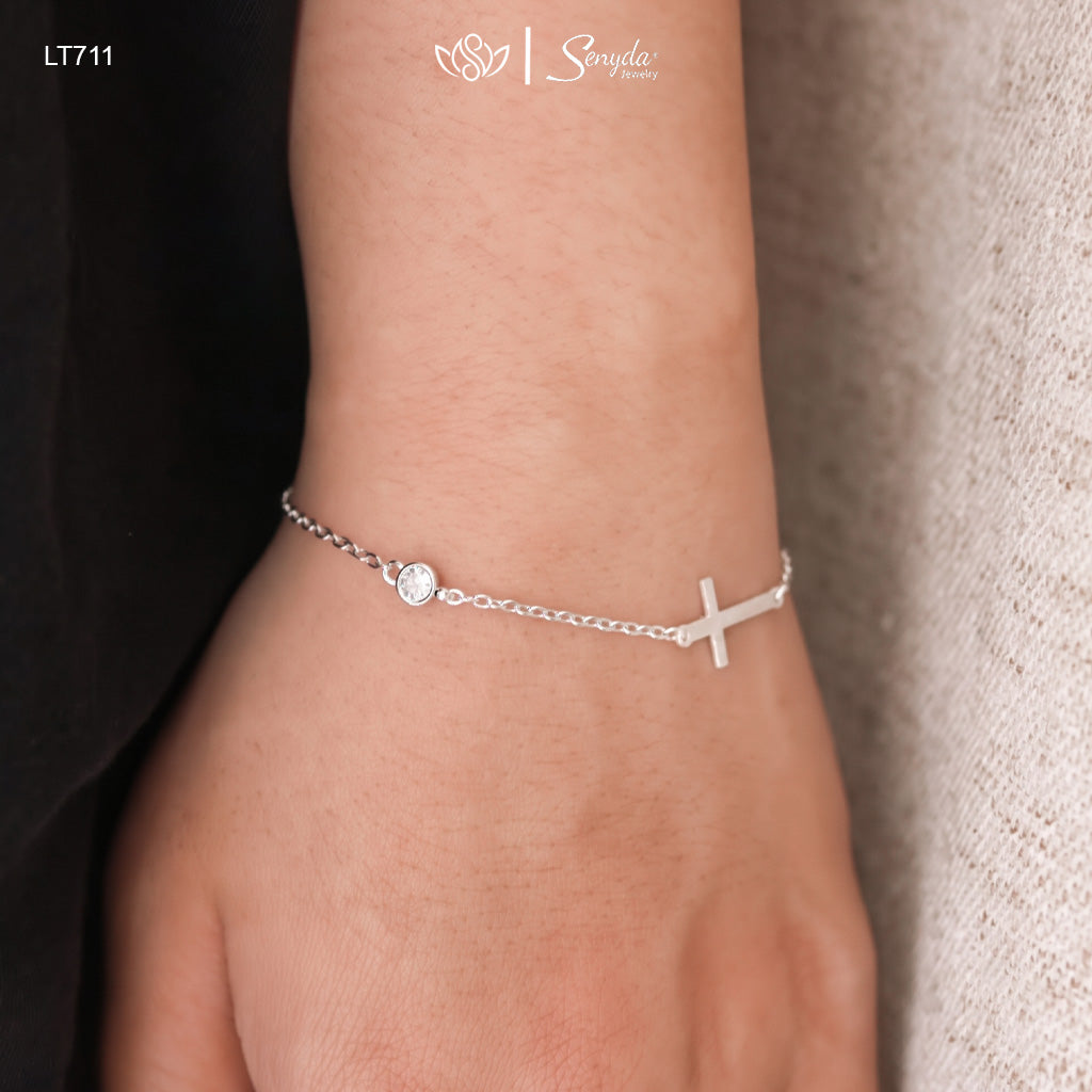 A close-up of a person's wrist wearing a delicate silver chain bracelet with a small cross and a round clear stone charm. The background shows a dark sleeve on the left and a textured light beige fabric on the right.