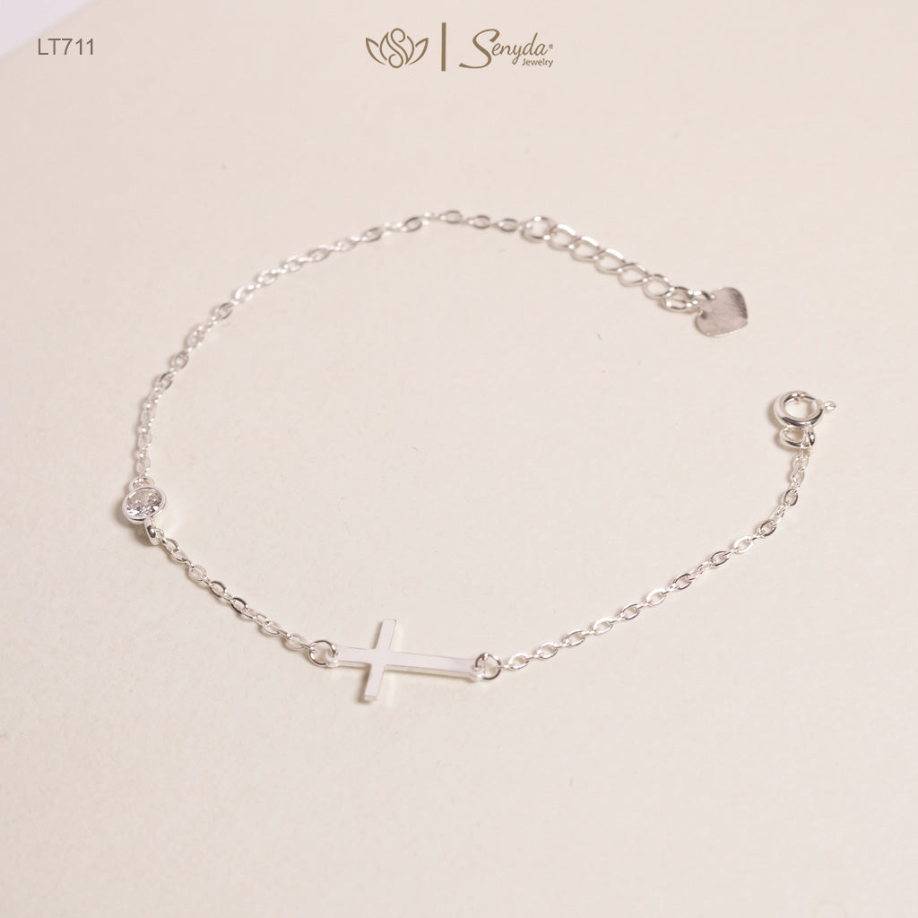 A delicate silver chain bracelet featuring a small cross charm in the center, a clear rhinestone on one side, and a small heart-shaped charm on the other, displayed on a light beige background with the Sonyda brand logo.