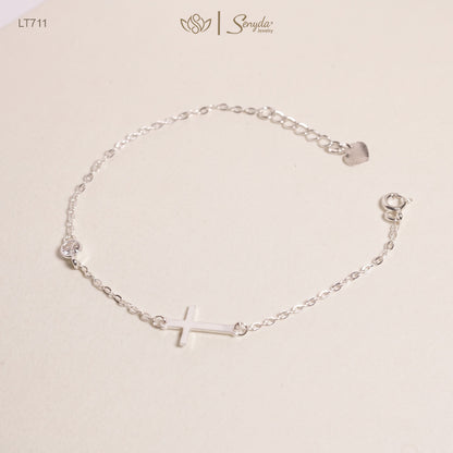 A delicate silver chain bracelet featuring a small cross charm in the center, a clear rhinestone on one side, and a small heart-shaped charm on the other, displayed on a light beige background with the Sonyda brand logo.