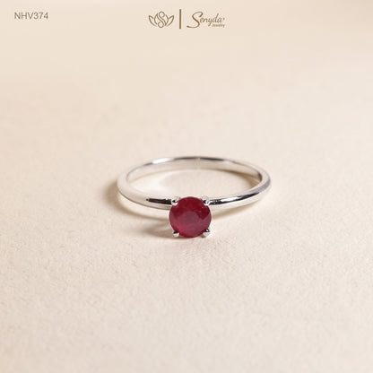 Dainty solid gold solitaire ring with a round red ruby in a four-prong setting on a cream background.