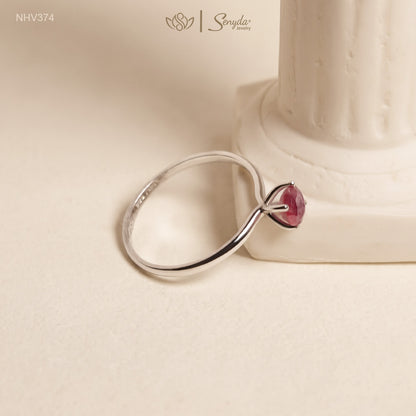 Dainty solid gold solitaire ring with a round red ruby in a four-prong setting on a cream background.